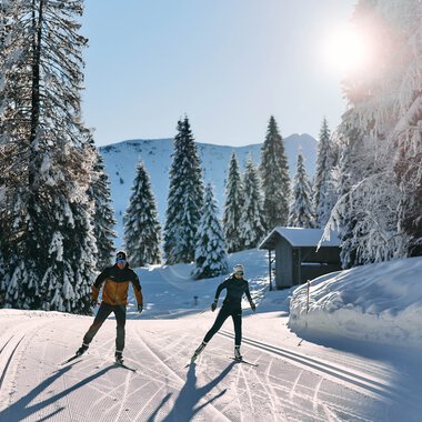 Zwei Skifahrer ziehen ihre Spuren im verschneiten Winterwald beim Langlaufen im Kleinwalsertal, Sonnenschein und klare Sicht. | ©  Kleinwalsertal Tourismus eGen • Fotograf: Oliver Farys