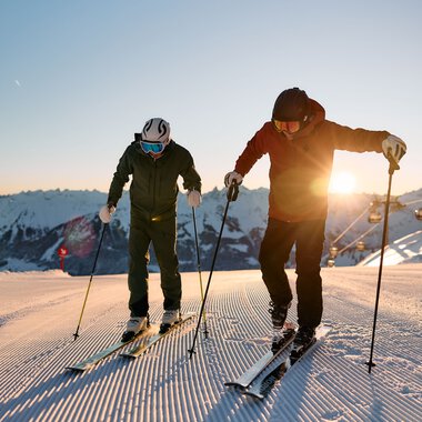 Zwei Skifahrer bei Sonnenaufgang auf einer verschneiten Piste im Kleinwalsertal, Alpen, beim Skifahren im Winter 2024. | © Kleinwalsertal Tourismus eGen • Fotograf: Oliver Farys