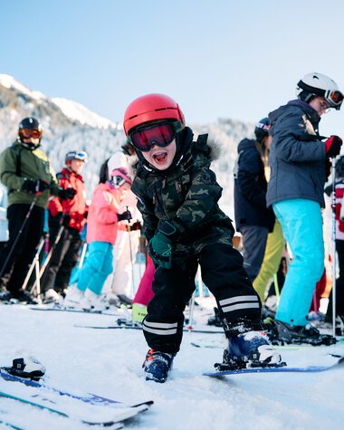 A young boy skiing and smiling, surrounded by other skiers in a snowy mountain landscape. | © Kleinwalsertal Tourismus eGen • Photographer: Oliver Farys