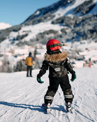 A child skier in winter clothing on a snowy slope, with mountains and other skiers in the background. | © Kleinwalsertal Tourismus eGen • Photographer: Oliver Farys