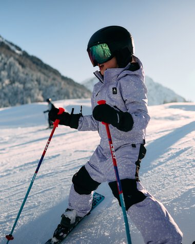 Child skiing in the snowy landscape, sunlight, mountains in the background, wearing helmet and ski gear. | © Kleinwalsertal Tourismus eGen • Photographer: Oliver Farys