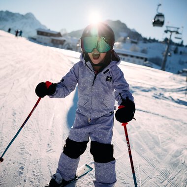 A young child skiing on a slope at a ski resort in Kleinwalsertal, surrounded by mountains, wearing goggles and winter clothing, holding ski poles. | © Kleinwalsertal Tourismus eGen • Photographer: Oliver Farys