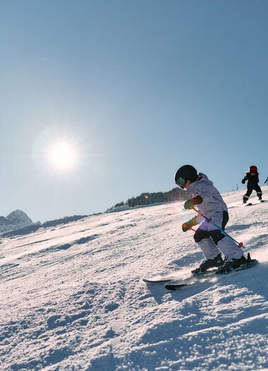 Child skiing on a snowy slope on a sunny day, with other children in the background and mountains in the distance. | © Kleinwalsertal Tourismus eGen • Photographer: Oliver Farys