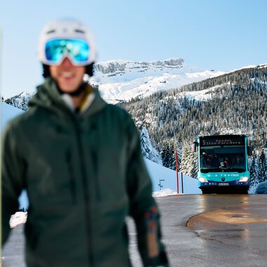 Person with skiing gear and helmet standing in front of snowy mountains in Kleinwalsertal, with a ski bus approaching in the background. | © Kleinwalsertal Tourismus eGen • Photographer: Oliver Farys