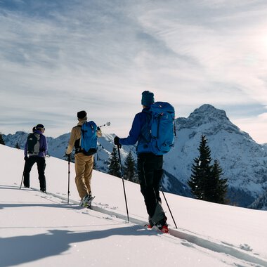 Three skitourers ascending in snowy mountain landscape in Kleinwalsertal, surrounded by mountains and pine trees, sunny winter weather. | © Kleinwalsertal Tourismus eGen • Photographer: Oliver Farys