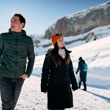 Two people walking in the snow-covered mountains under a clear blue sky, with a rugged landscape in the background. | © Kleinwalsertal Tourismus eGen • Photographer: Oliver Farys
