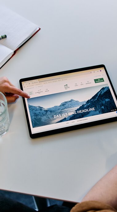 Vier Personen sitzen um einen Tisch und schauen auf ein Tablet mit einer Berglandschaft auf dem Bildschirm, im Büro während eines Meetings. | © Kleinwalsertal Tourismus eGen • Fotograf: Oliver Farys