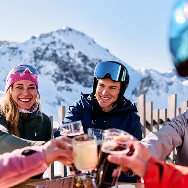 Group of friends at a ski lodge enjoying drinks and toasting, with snowy mountains in the background. | © Kleinwalsertal Tourismus eGen • Photographer: Oliver Farys