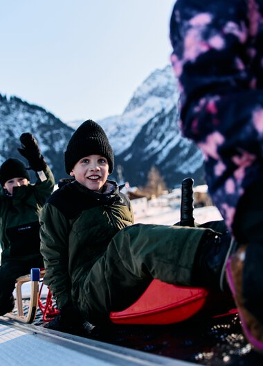 Children sledding in the snow in the mountains, laughing and playing, winter vacation in Kleinwalsertal. | © Kleinwalsertal Tourismus eGen • Photographer: Oliver Farys