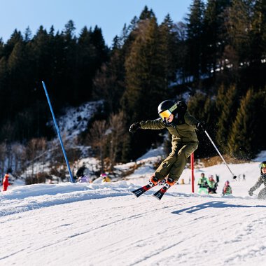 Children skiing on a snow-covered slope in Kleinwalsertal, surrounded by trees and winter scenery, in sunny weather. | © Kleinwalsertal Tourismus eGen • Photographer: Oliver Farys