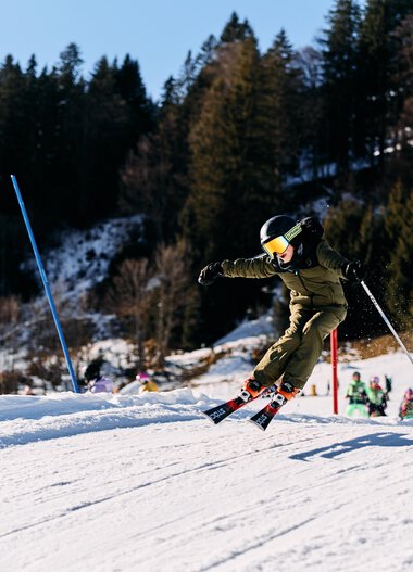 Kinder beim Skifahren auf einer Skipiste im Kleinwalsertal, umgeben von Bäumen und winterlicher Landschaft, bei Sonnenschein. | © Kleinwalsertal Tourismus eGen • Fotograf: Oliver Farys