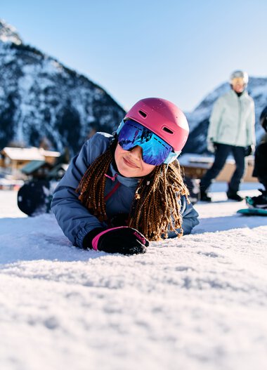 Girl lying on snow in a ski area, wearing a pink helmet and ski goggles, with mountains and other skiers in the background. | © Kleinwalsertal Tourismus eGen • Photographer: Oliver Farys