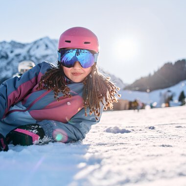 Girl skiing in the mountains, wearing a pink helmet and ski goggles, lying in the snow with snow-covered mountains in the background. | © Kleinwalsertal Tourismus eGen • Photographer: Oliver Farys