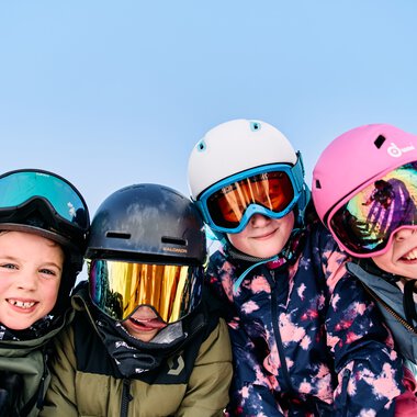 Four children in winter clothing and helmets, smiling and posing in the snow while skiing. | © Kleinwalsertal Tourismus eGen • Photographer: Oliver Farys