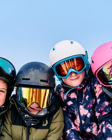 Four children in winter clothing and helmets, smiling and posing in the snow while skiing. | © Kleinwalsertal Tourismus eGen • Photographer: Oliver Farys