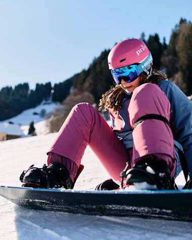 Young female skier in pink jacket and helmet sitting on her snowboard in the snow, with a winter landscape featuring snow-covered mountains and trees in the background. | © Kleinwalsertal Tourismus eGen • Photographer: Oliver Farys