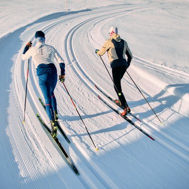 Two cross-country skiers in classic style on a snow-covered trail in Kleinwalsertal, Schwarzwasserloipe, during the winter 2025 season. | © Kleinwalsertal Tourismus eGen • Photographer: Oliver Farys