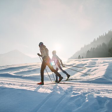Two cross-country skiers on the Schwarzwasserloipe in Kleinwalsertal during sunrise, surrounded by snowy landscape and mountains. | © Kleinwalsertal Tourismus eGen • Photographer: Oliver Farys
