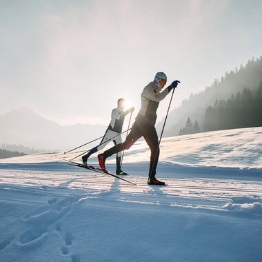 Two cross-country skiers in a snowy landscape with the sun in the background, on a trail in Kleinwalsertal. | © Kleinwalsertal Tourismus eGen • Photographer: Oliver Farys