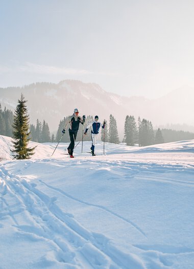 Two people cross-country skiing in a snowy landscape with a wooden cabin and pine trees in the background. | © Kleinwalsertal Tourismus eGen • Photographer: Oliver Farys