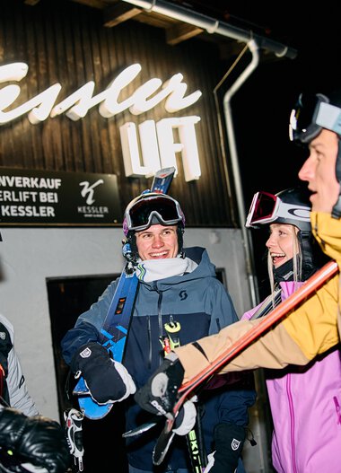 Four skiers in winter gear with helmets and skis, happily socializing at night in Kleinwalsertal, with a neon sign glowing in the background. | © Kleinwalsertal Tourismus eGen • Photographer: Oliver Farys