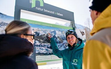 Woman in winter clothing points at a large information board with mountain images while two other people listen attentively, in snowy Kleinwalsertal. | © Kleinwalsertal Tourismus eGen • Photographer: Oliver Farys