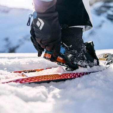 Person beim Schneeschuhwandern im Schnee, mit einer Hand, die eine Kamera hält, im Hintergrund verschneite Berge. | © Kleinwalsertal Tourismus eGen • Fotograf: Oliver Farys