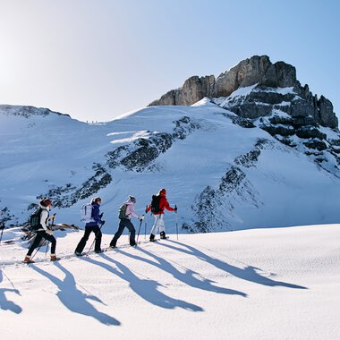 Sechs Personen beim Schneeschuhwandern in verschneiter Berglandschaft im Kleinwalsertal, Sonne am Himmel. | © Kleinwalsertal Tourismus eGen • Fotograf: Oliver Farys