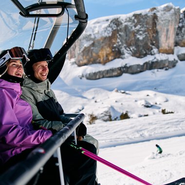 Two people sitting on a ski lift in winter in Kleinwalsertal, surrounded by snowy landscape and mountains. | © Kleinwalsertal Tourismus eGen • Photographer: Oliver Farys