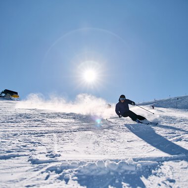 Skifahrer am Kanzelwand in Kleinwalsertal bei strahlendem Sonnenschein, im Hintergrund Skilifte und eine Berghütte. | © Kleinwalsertal Tourismus eGen • Fotograf: Oliver Farys