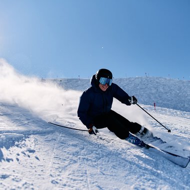 Skier slalom down a snowy slope in Kanzelwand, with a clear blue sky and bright sun in the background, wearing a helmet and dark clothing. | © Kleinwalsertal Tourismus eGen • Photographer: Oliver Farys