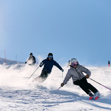 Drei Skifahrer in Aktion auf einer verschneiten Piste im Kleinwalsertal, mit Bergpanorama im Hintergrund, bei schönem Winterwetter. | © Kleinwalsertal Tourismus eGen • Fotograf: Oliver Farys