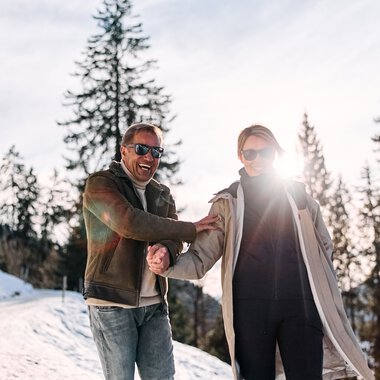 Two smiling people on a winter hike in Kleinwalsertal, shaking hands warmly, surrounded by snow-covered trees. | © Kleinwalsertal Tourismus eGen • Photographer: Oliver Farys