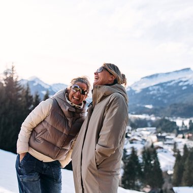 Two laughing women enjoying a winter hike in a snowy landscape with mountains in the background. | © Kleinwalsertal Tourismus eGen • Photographer: Oliver Farys