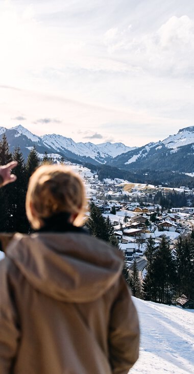 Zwei Personen schauen auf eine winterliche Berg-/Tal-Landschaft vom Schnee bedeckten Hügel aus, mit Häusern, Wäldern und Bergen im Hintergrund. | © Kleinwalsertal Tourismus eGen • Fotograf: Oliver Farys