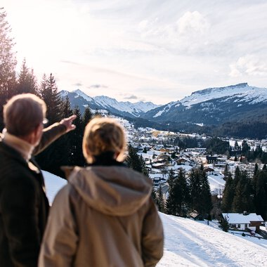 Two people overlook a snowy mountain and valley landscape with houses, forests, and mountains in the background from a snow-covered hill. | © Kleinwalsertal Tourismus eGen • Photographer: Oliver Farys