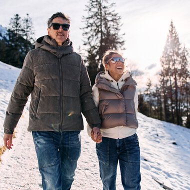 A man and a woman walking in the snowy winter landscape, surrounded by pine trees, under sunny weather, dressed warmly. | © Kleinwalsertal Tourismus eGen • Photographer: Oliver Farys