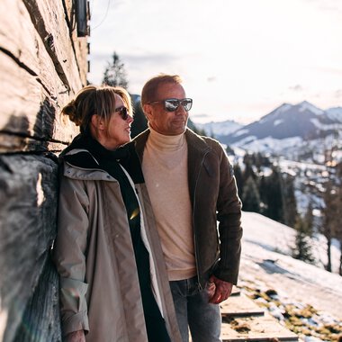 Two people standing during a winter hike in the snow, with mountains in the background, next to a wooden wall on a sunny day. | © Kleinwalsertal Tourismus eGen • Photographer: Oliver Farys