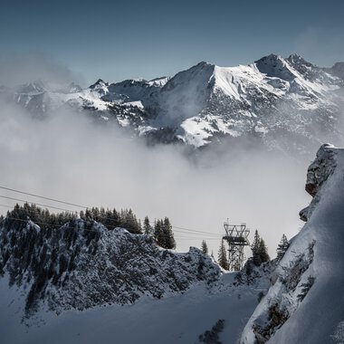 Schneebedeckte Berge und Nebel in einer Winterlandschaft mit Wald im Vordergrund. | © Kleinwalsertal Tourismus eGen • Fotograf: Robert Niederwolfsgruber