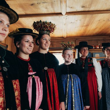 Sechs Frauen in traditioneller bayerischer Tracht stehen nebeneinander in einem Holzraum, lächeln und schauen in eine Richtung. | © Kleinwalsertal Tourismus eGen • Fotograf: Stefan Klauser 