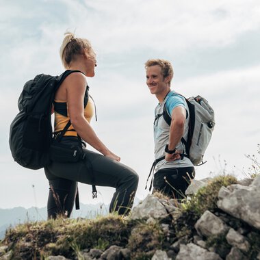 Two hikers on a mountain trail, standing on a rocky path with mountain views in the background. | © Kleinwalsertal Tourismus eGen • Photographer: Stefan Klauser