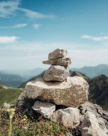 Steinmännchen auf einer Bergkuppe mit Blick auf grüne Hügel und Berge im Hintergrund, blauer Himmel mit wenigen Wolken. | © Kleinwalsertal Tourismus eGen • Fotograf: Stefan Klauser 