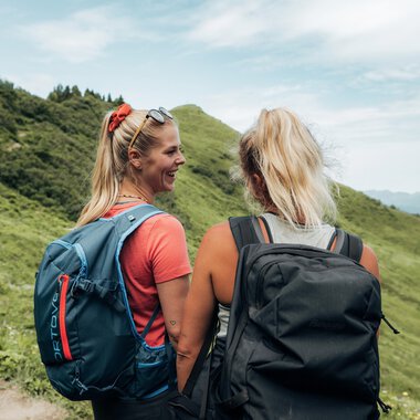 Two women with backpacks hiking in the mountains, with green hills and a blue sky in the background. | © Kleinwalsertal Tourismus eGen • Photographer: Stefan Klauser