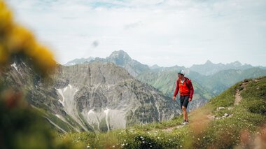 Ein Wanderer mit Rucksack und Hut unterwegs in den Bergen, umgeben von grünen Wiesen und hohen Gipfeln, im Hintergrund bewölkter Himmel. | © Kleinwalsertal Tourismus eGen • Fotograf: Stefan Klauser 