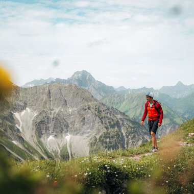 Ein Wanderer mit Rucksack und Hut unterwegs in den Bergen, umgeben von grünen Wiesen und hohen Gipfeln, im Hintergrund bewölkter Himmel. | © Kleinwalsertal Tourismus eGen • Fotograf: Stefan Klauser 