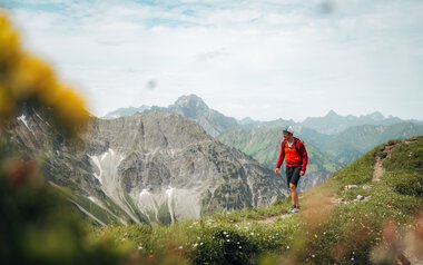 A hiker with a backpack and hat walking in the mountains, surrounded by green meadows and high peaks, with a cloudy sky in the background. | © Kleinwalsertal Tourismus eGen • Photographer: Stefan Klauser