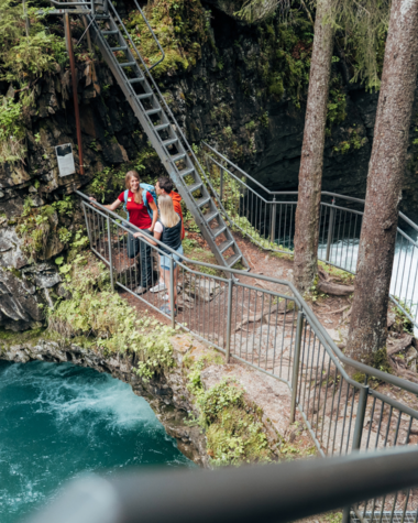 Zwei Personen in roter Kleidung auf einem Holzsteg im Wald neben einem Wasserfall, umgeben von grünen Bäumen. | ©  Kleinwalsertal Tourismus eGen • Fotograf: Stefan Klauser 