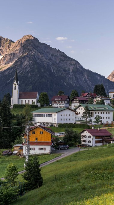 Panoramaaufnahme eines kleinen Dorfes in den Bergen mit Kirche, umgeben von grünen Wiesen und Bäumen, vor einer dramatischen Bergkulisse. | © Kleinwalsertal Tourismus eGen • Fotograf: Steffen Berschin 