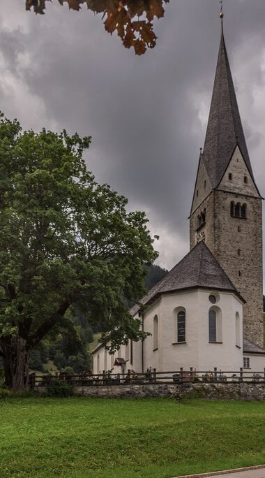 Ein Kirchengebäude mit einem hohen Turm, umgeben von Bäumen und einem Wohnhaus. | © Kleinwalsertal Tourismus eGen • Fotograf: Steffen Berschin 