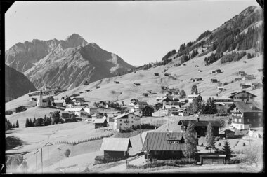 Historisches Schwarz-Weiß-Foto eines kleinen Dorfes in den österreichischen Alpen mit Kirche, umgeben von Bergen und Hütten. | © Kleinwalsertal Tourismus eGen • Karl Max Kessler Archiv